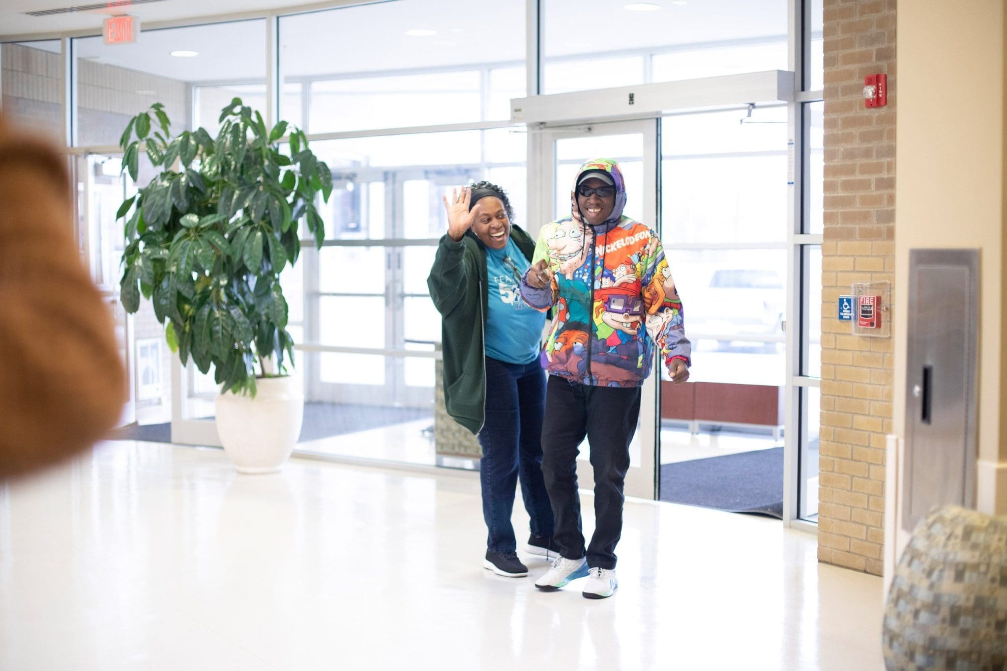 Young man with Autism Spectrum Disorder (ASD) in a brightly colored jacket grins as he walks alongside his smiling mother after a therapy appointment, feeling accomplished and supported.