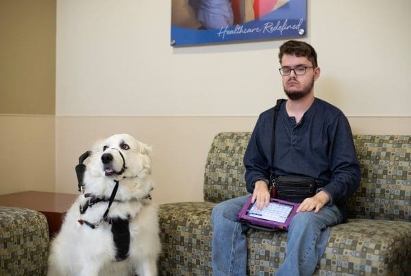 A young man and his service dog wait in the office of a clinic for those with Intellectual and developmental disabilities. He has his Augmentative and Alternative Communication device in his lap.